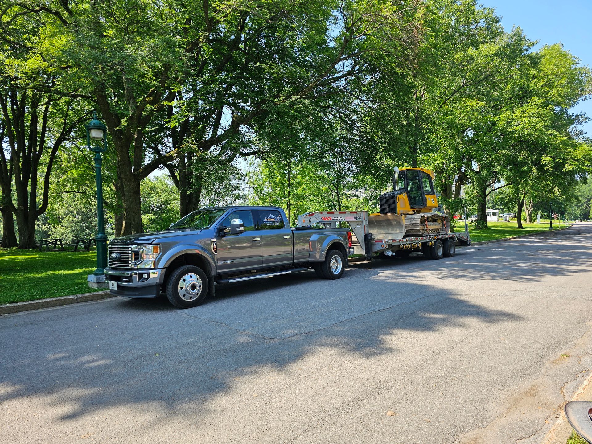 Une camionnette argentée tractant une remorque avec une pelleteuse jaune dans une rue bordée d'arbres