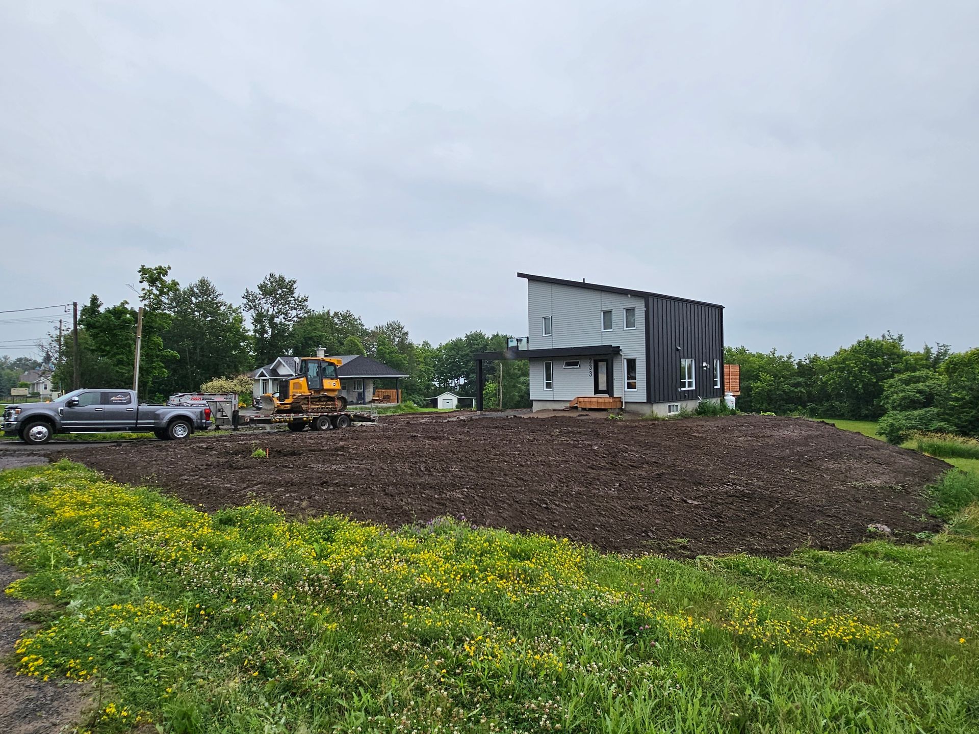 Chantier de construction avec excavatrice et camionnette à côté d'une petite maison grise sur une colline de terre sous un ciel nuageux.