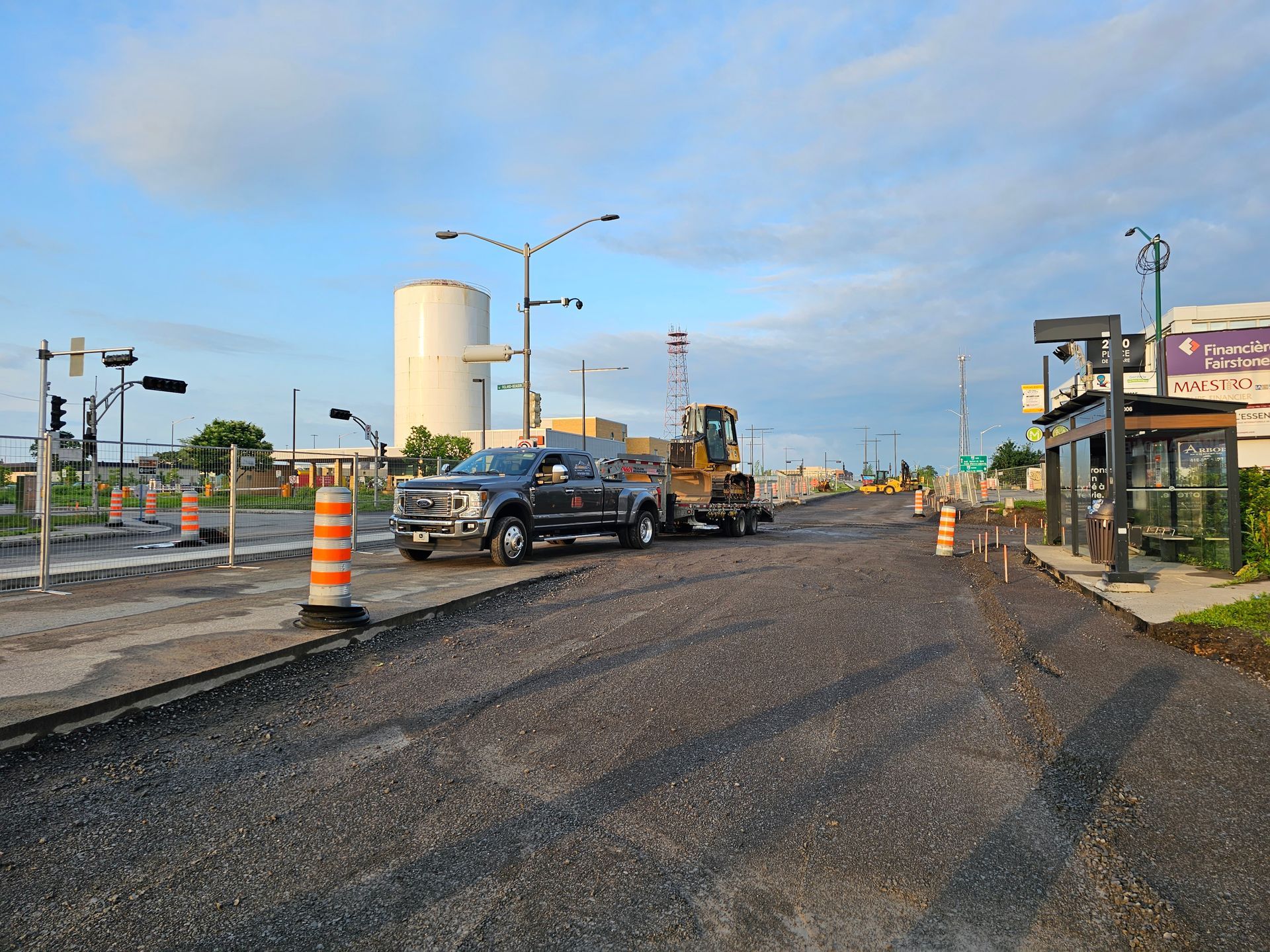 Zone de travaux sur une route de gravier avec des camions, des cônes orange et des lampadaires sous un ciel bleu