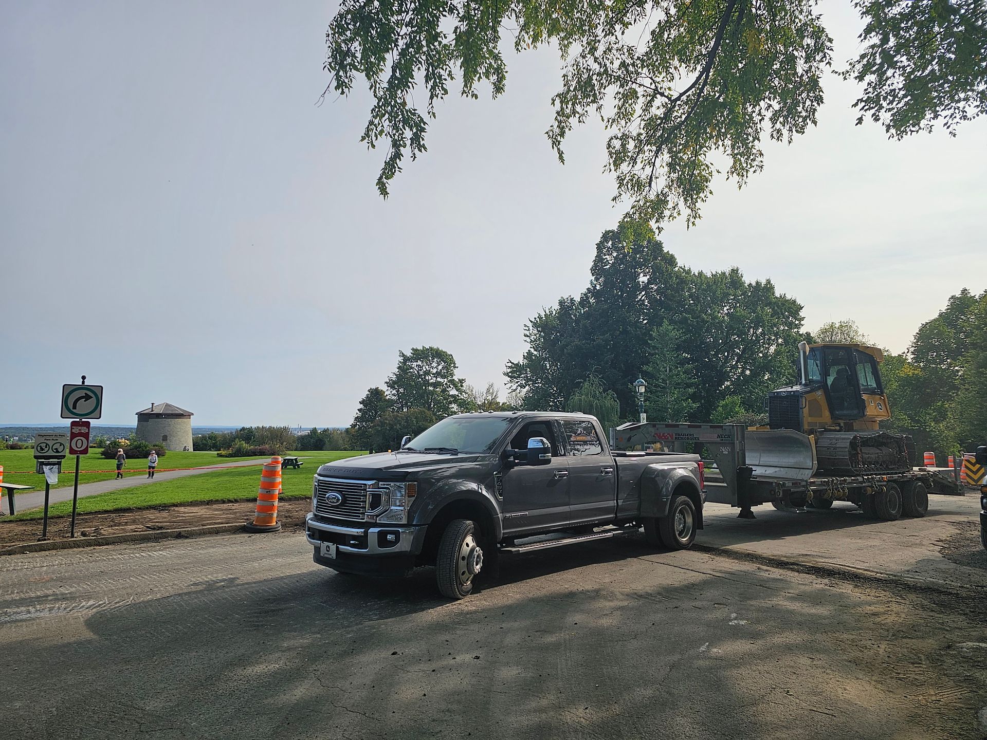 Une camionnette noire tracte de l'équipement lourd sur un chemin de gravier, sur un chantier, avec des arbres et un point d'eau à proximité.