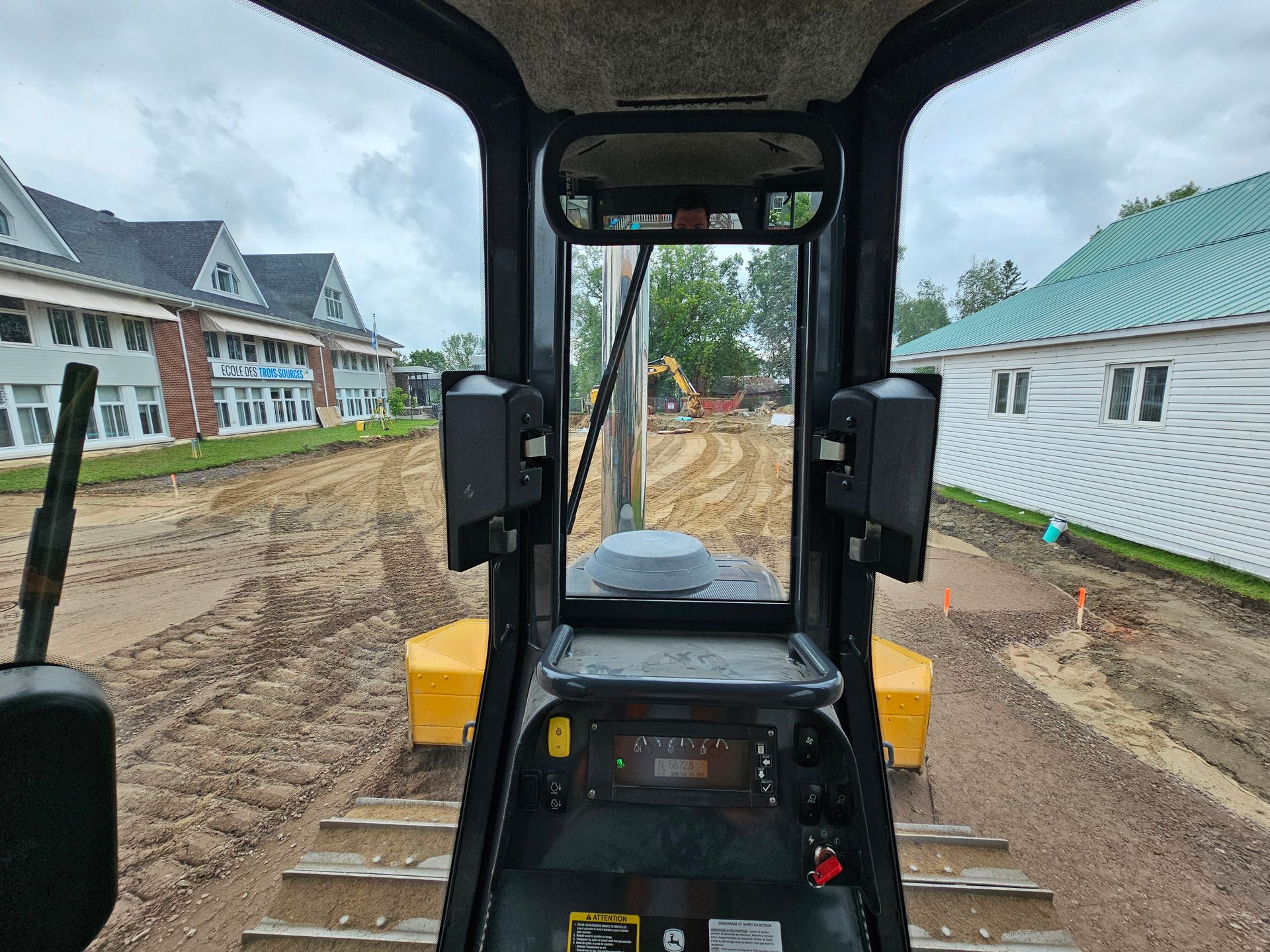 Vue depuis la cabine d'un engin de chantier sur un chantier boueux bordé de maisons de part et d'autre.