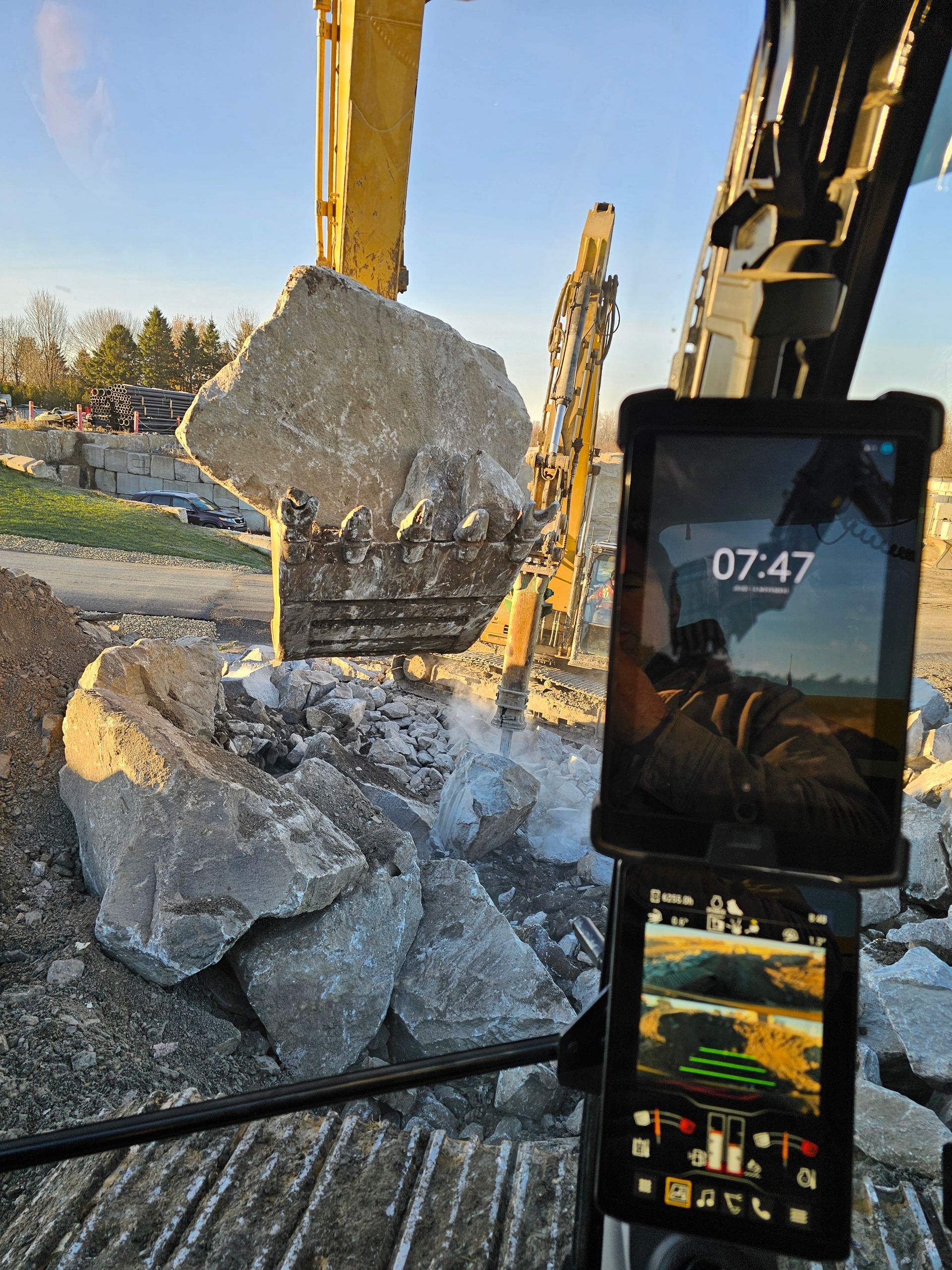 Le godet d'une excavatrice soulève un gros rocher sur un chantier de construction, avec un écran de téléphone visible au premier plan.