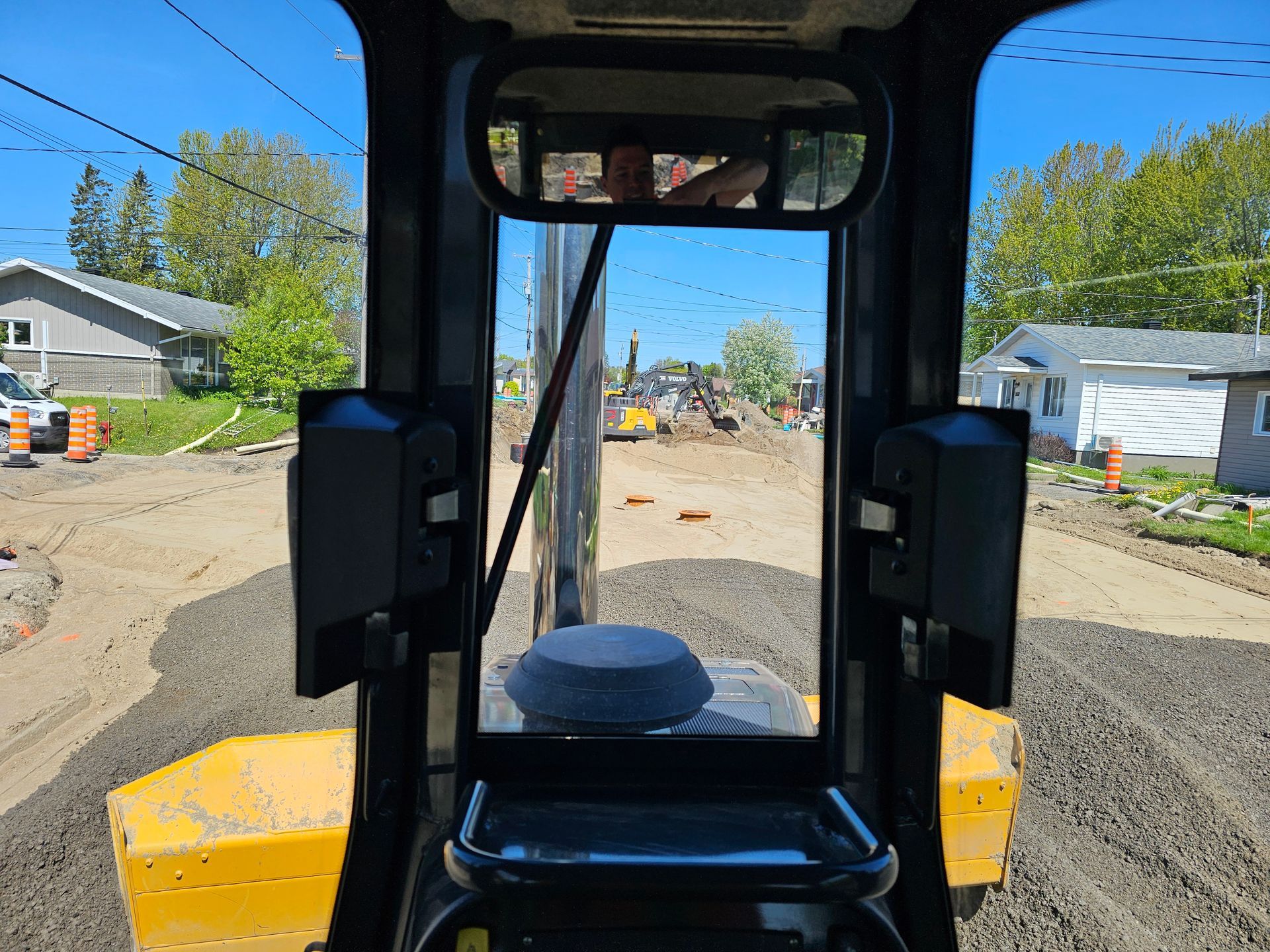 À l'intérieur de la cabine d'un engin de chantier, vue sur une route de gravier et des maisons par une journée ensoleillée.