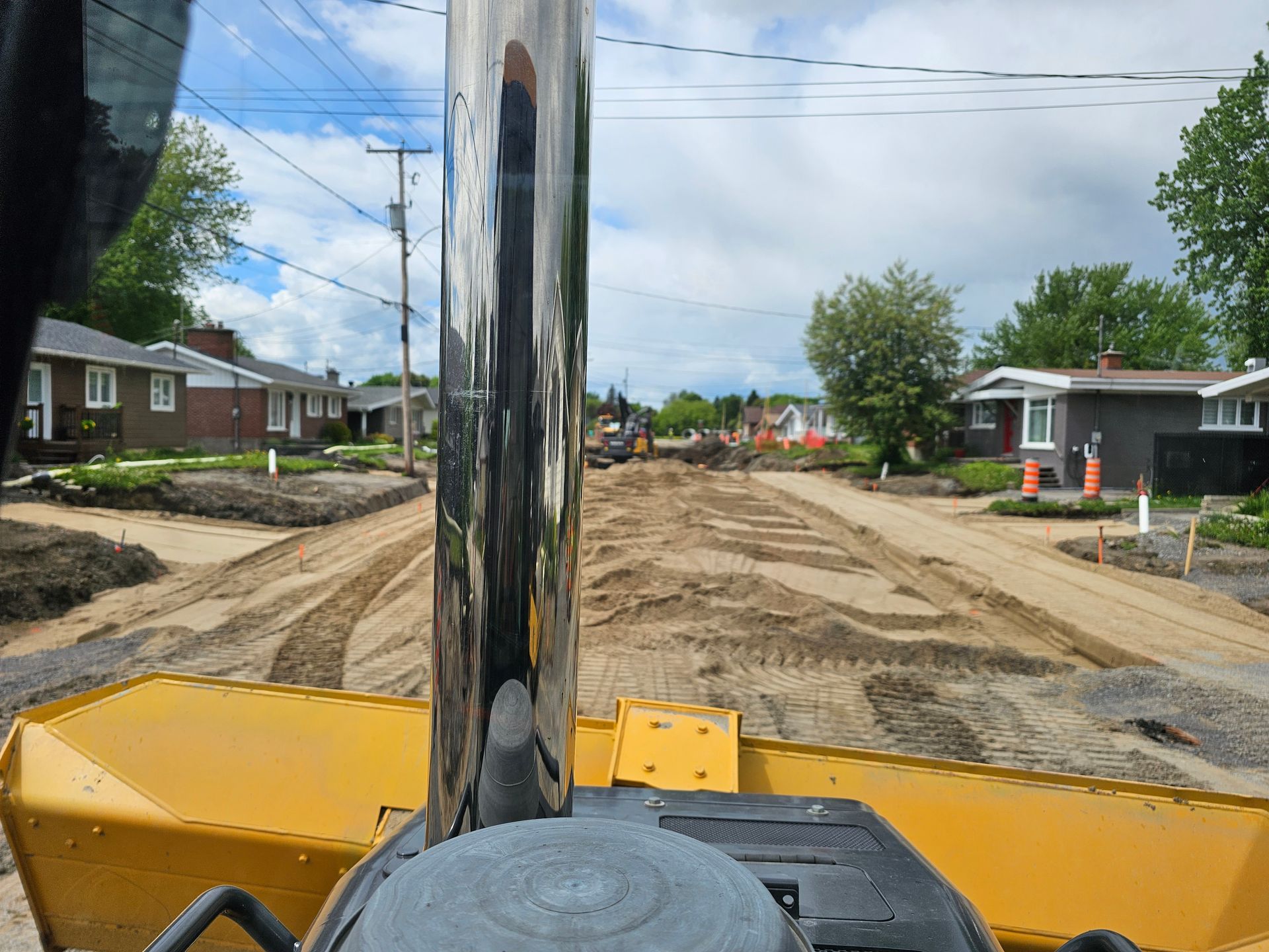 Vue d'un bulldozer sur une rue de terre bordée de maisons et de poteaux électriques sous un ciel nuageux