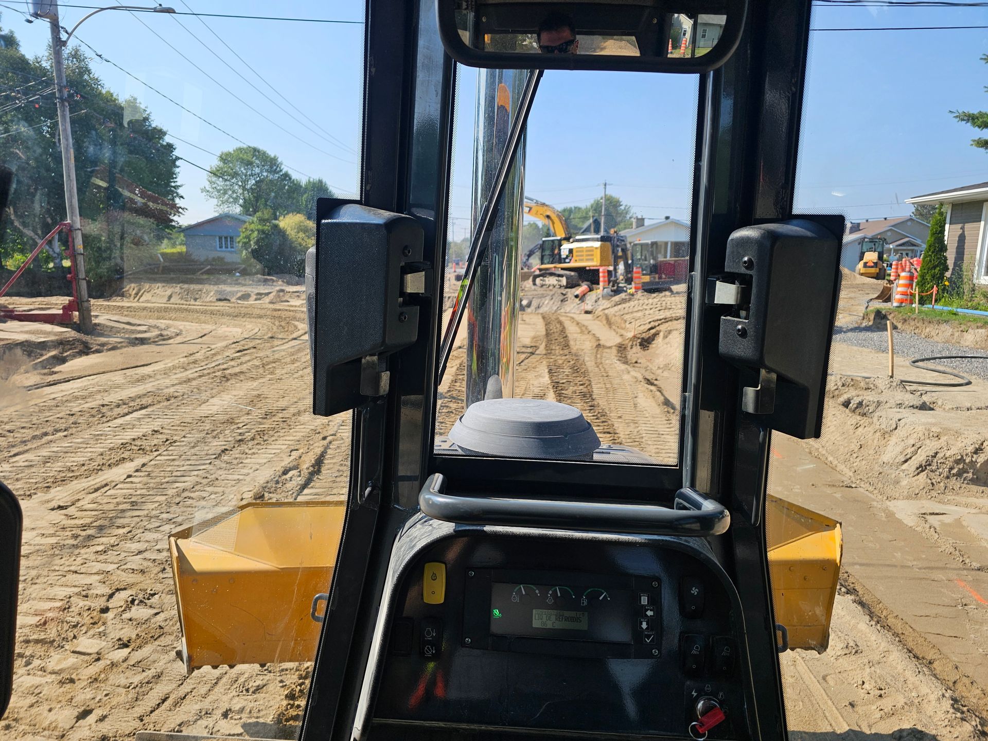 Vue depuis la cabine d'une excavatrice jaune sur un chantier de construction sablonneux avec des ouvriers et des engins devant.