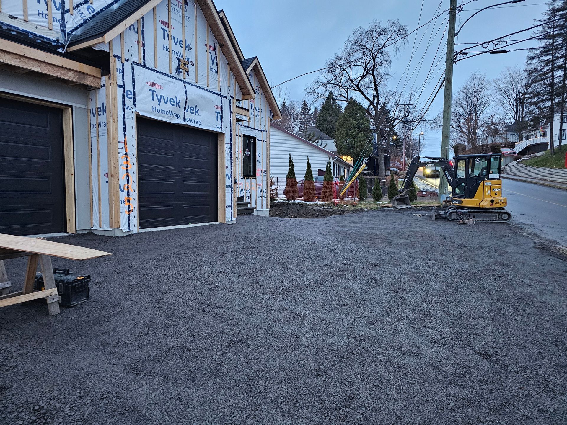 Allée de gravier longeant des garages blancs aux portes noires, table de pique-nique et une maison éclairée au loin.