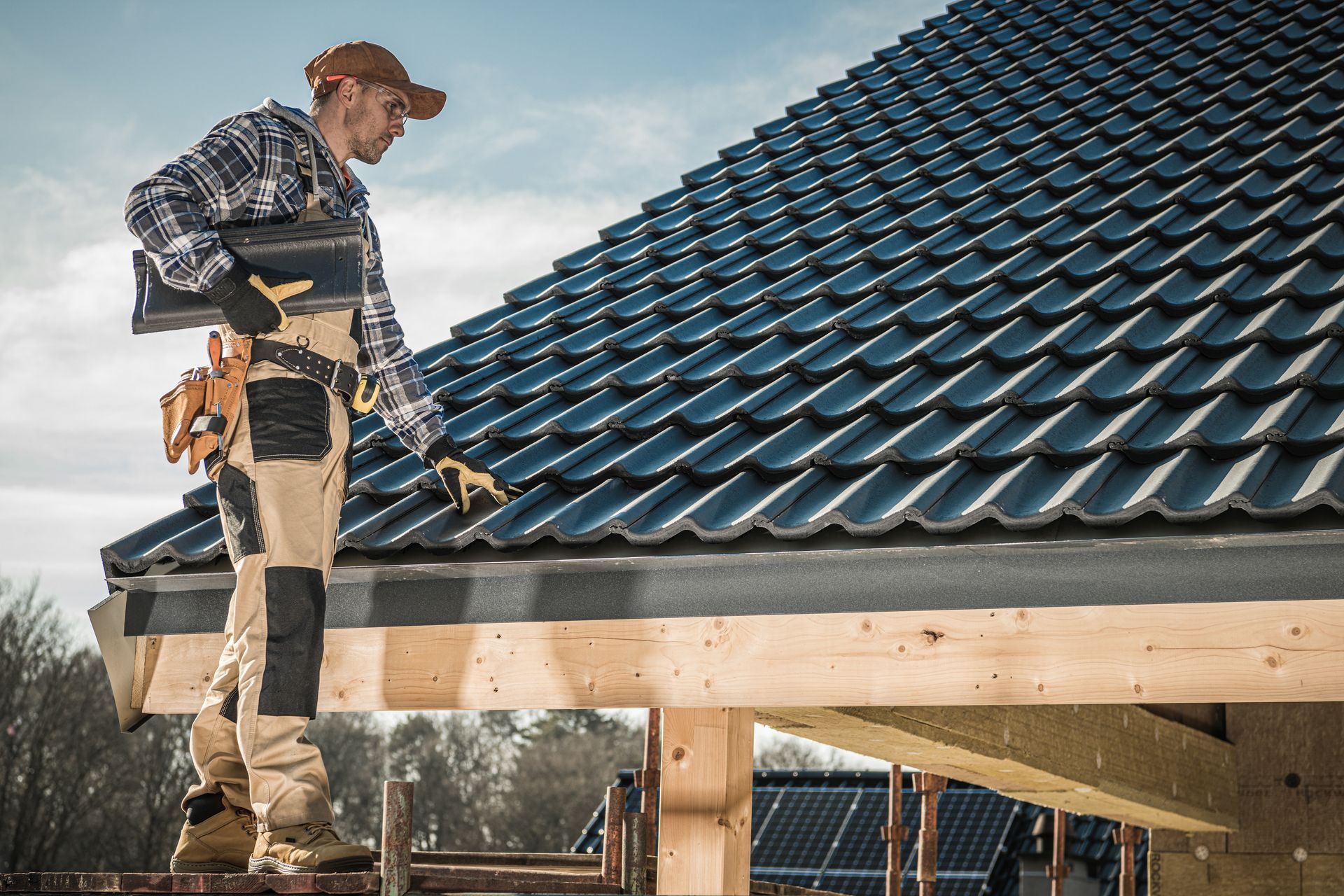 A male worker, standing on a ladder, holds roofing shingles as he stands next to a shingled roof.