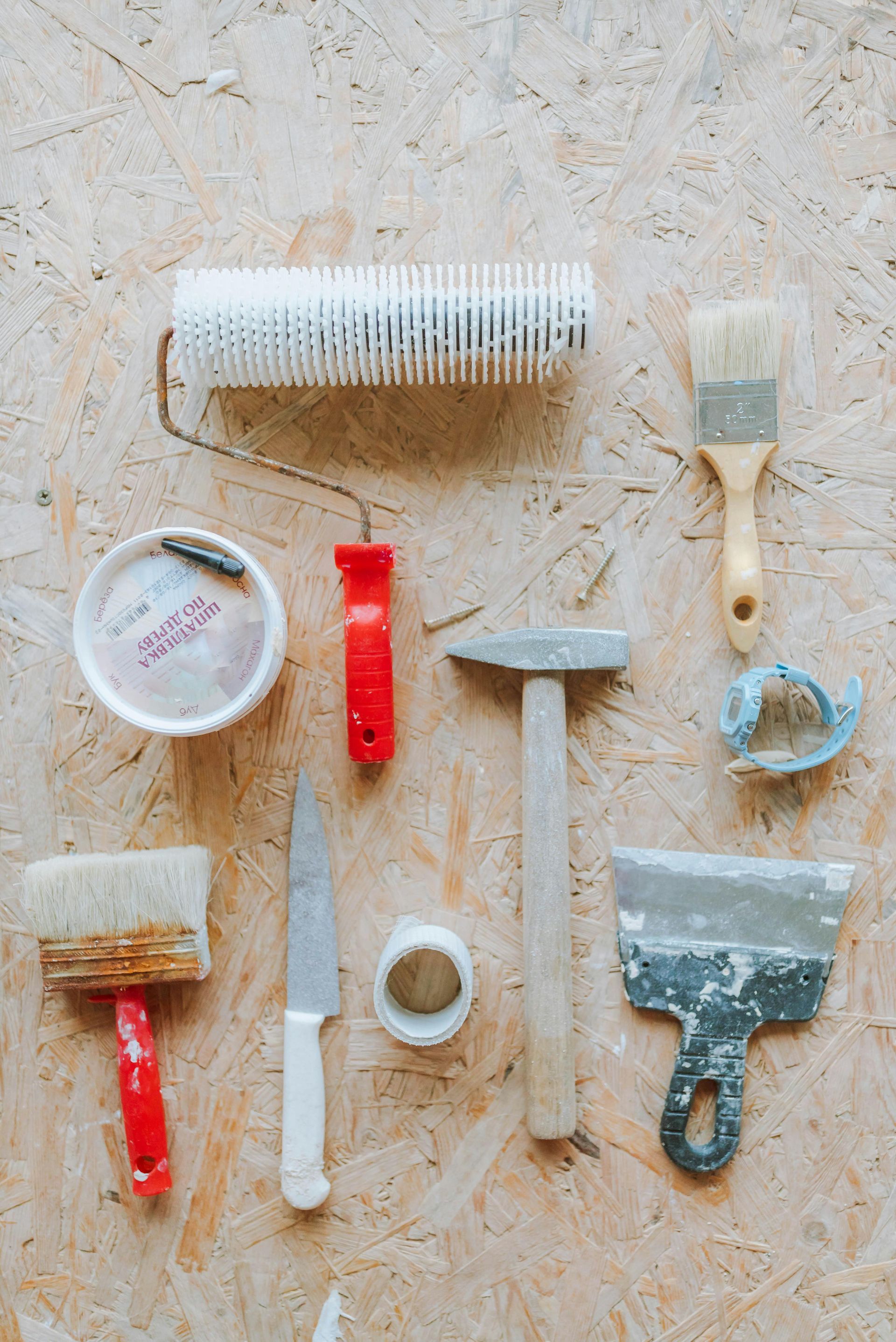 Assorted paint and wallpaper tools arranged on a textured floor, including brushes, roller, scraper, tray, and pliers.