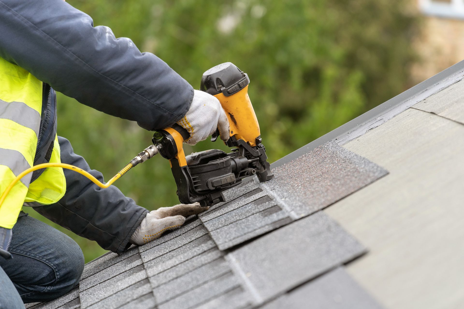 roofer worker in uniform work wear using air or pneumatic nail gun and installing asphalt or bitumen tile 