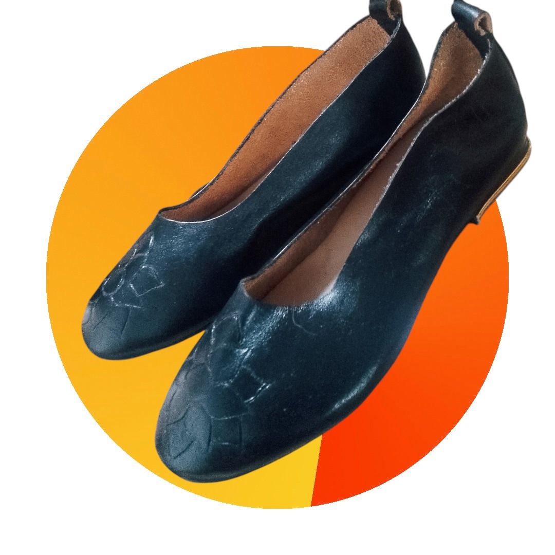 Un par de zapatos planos de piel negra con un sutil relieve floral en la puntera, sobre un fondo circular naranja.