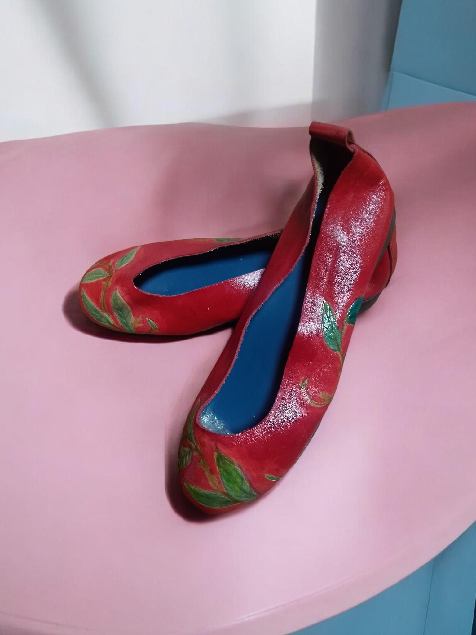 Un par de zapatos planos rojos con un estampado de hojas verdes y plantillas azules, sobre una superficie rosa.