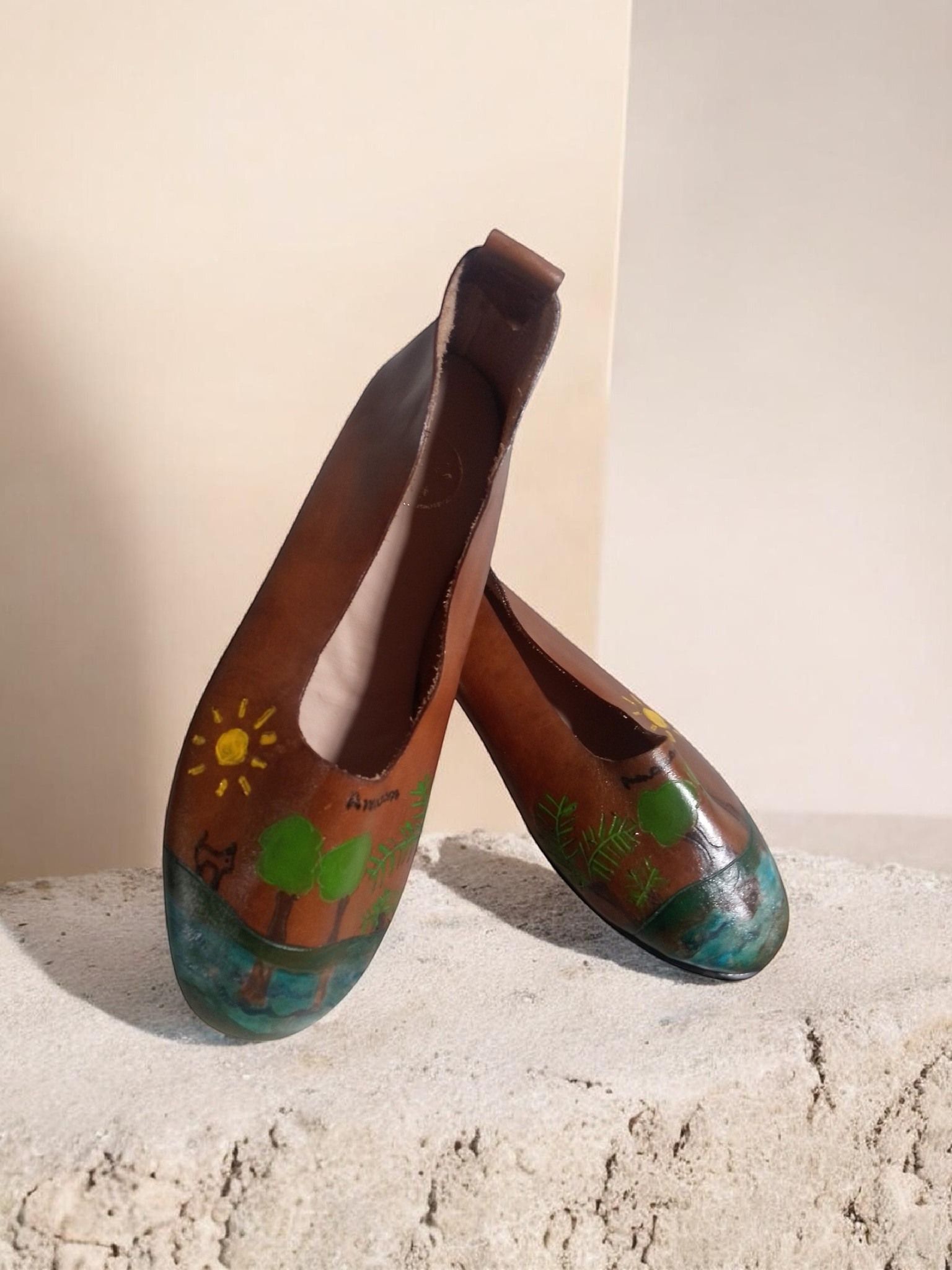 Un par de zapatos sin cordones de cuero marrón pintados a mano con un sol, árboles verdes 