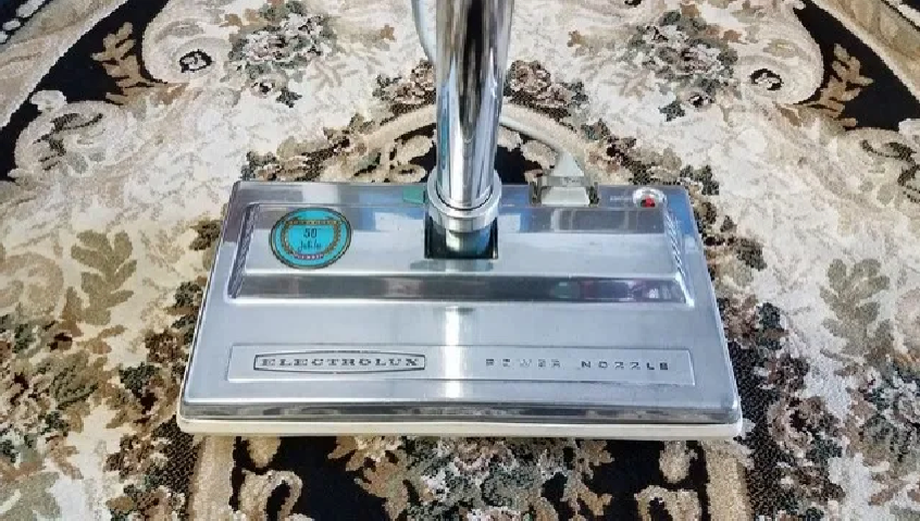 A shiny, metallic Electrolux vacuum floor nozzle sits centered on a dark patterned rug.