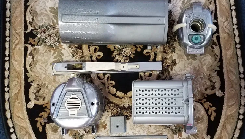 Disassembled metallic parts of a vintage projector, including a casing, lens housing, and internal components on a rug.