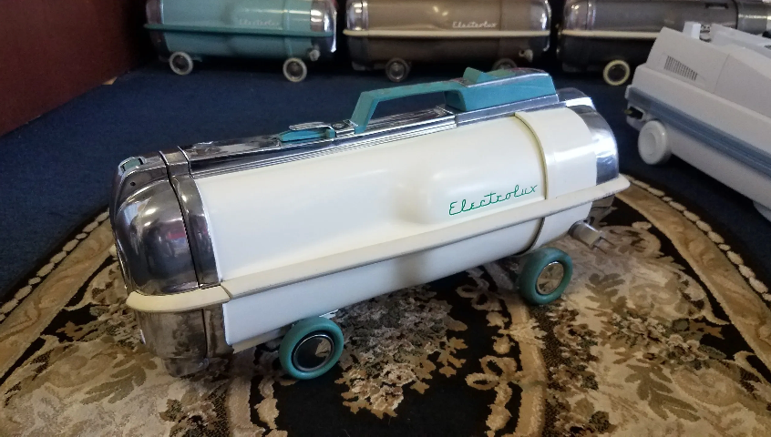 A vintage white and teal Electrolux canister vacuum cleaner sits on a patterned rug with other vacuums in the background.