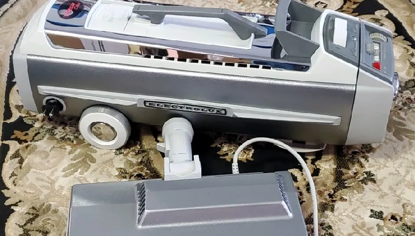 A silver and white vintage canister-style vacuum cleaner sits on a patterned rug.