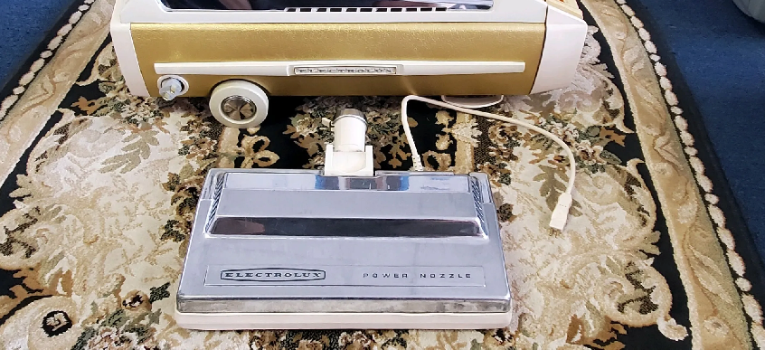 A gold-and-white vacuum cleaner unit with a chrome floor nozzle attachment resting on a patterned rug.