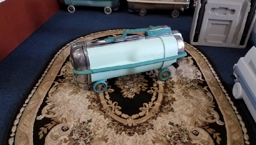 A light blue and chrome vintage canister vacuum cleaner sits on an ornate, brown and cream patterned area rug.