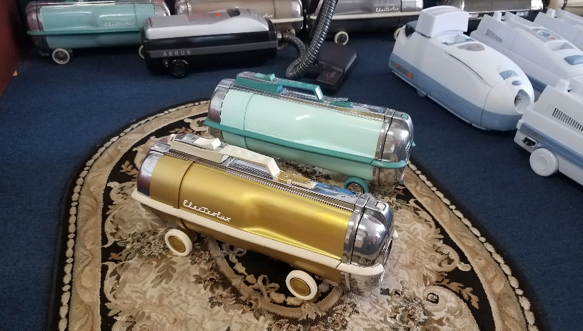 A collection of vintage canister vacuum cleaners, featuring gold and mint green models on a patterned rug.
