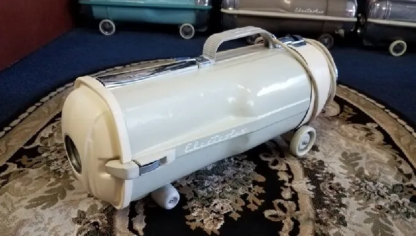 Vintage beige canister vacuum cleaner with chrome accents on wheels sitting on a patterned rug.