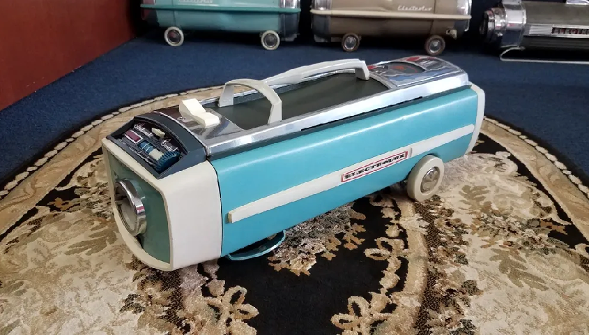 A turquoise and white vintage canister vacuum cleaner sits on a patterned rug, with other units visible in the background.