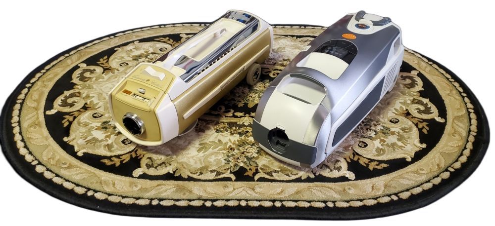 Two compact, handheld projectors, one gold and one silver, sit side-by-side on an oval, ornate patterned rug.