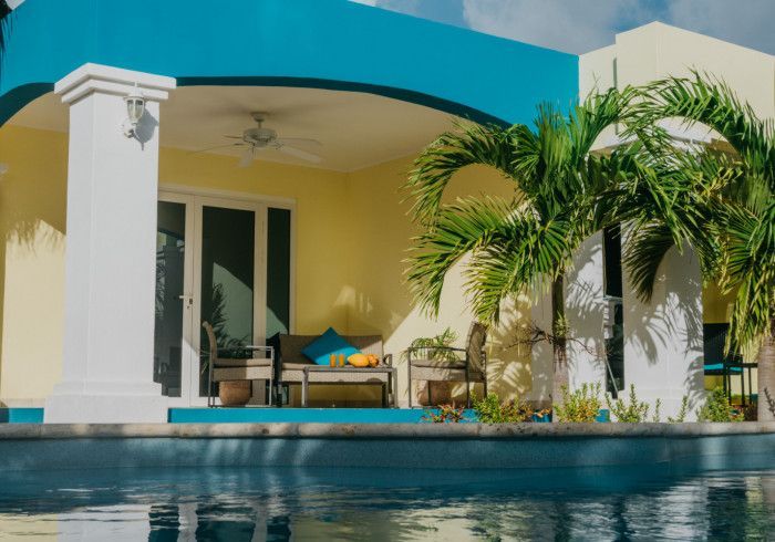 Poolside resort with a blue and yellow facade, palm trees, and a covered patio with seating.