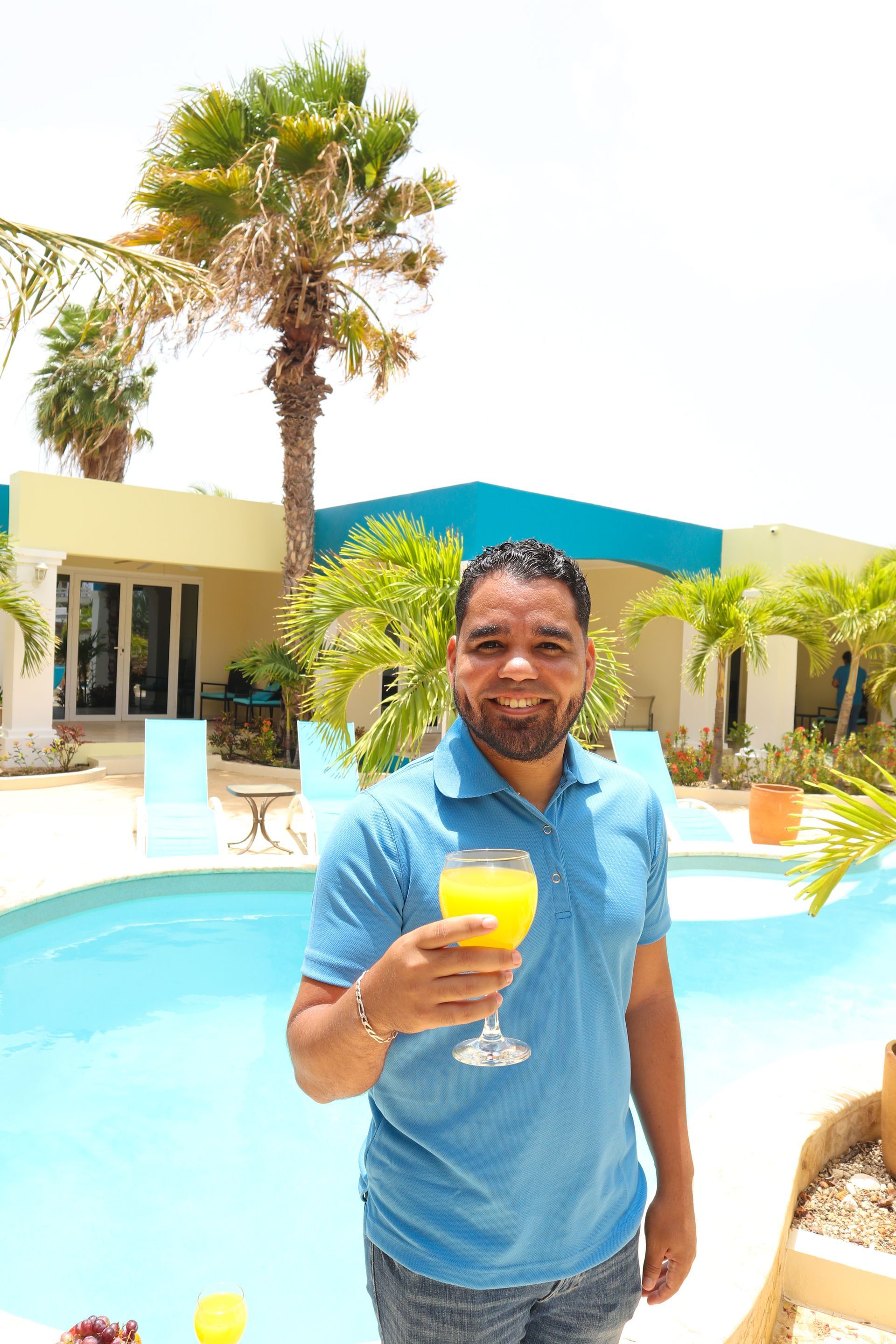 Man holding orange drink by a pool with a tropical backdrop.