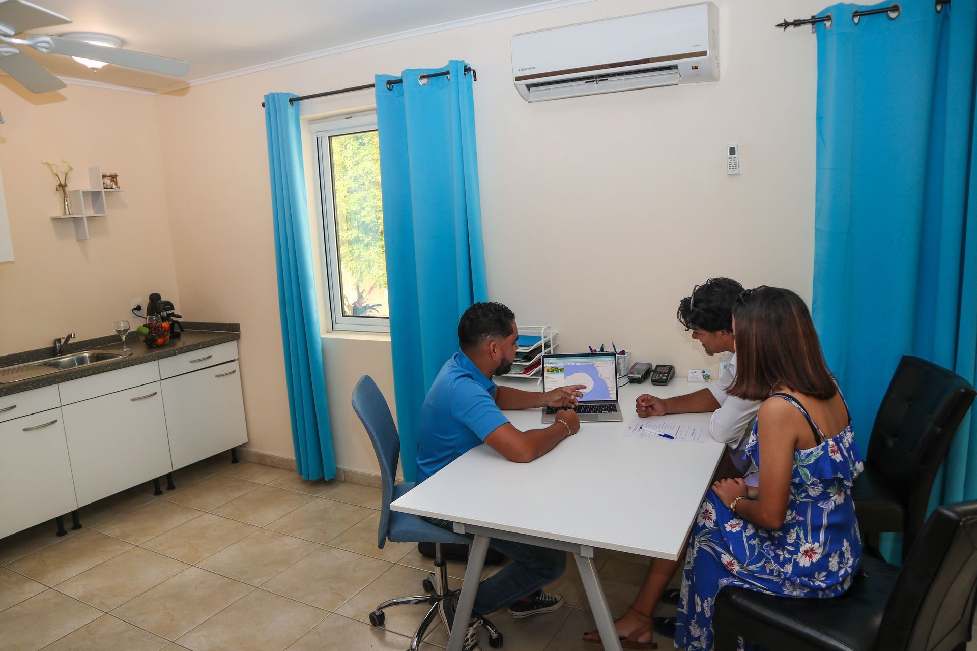 Three people at a white table in a room. One person shows a laptop screen. Blue curtains, AC unit, and white cabinets.