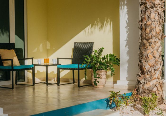 Patio with chairs, table, and potted plant, under a yellow wall, lit by sunlight.