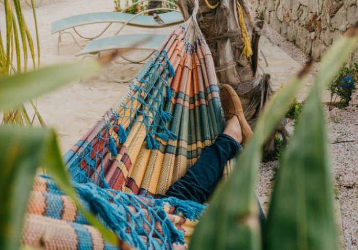 Person relaxing in a colorful hammock, feet visible. Palm tree in background, sandy setting.