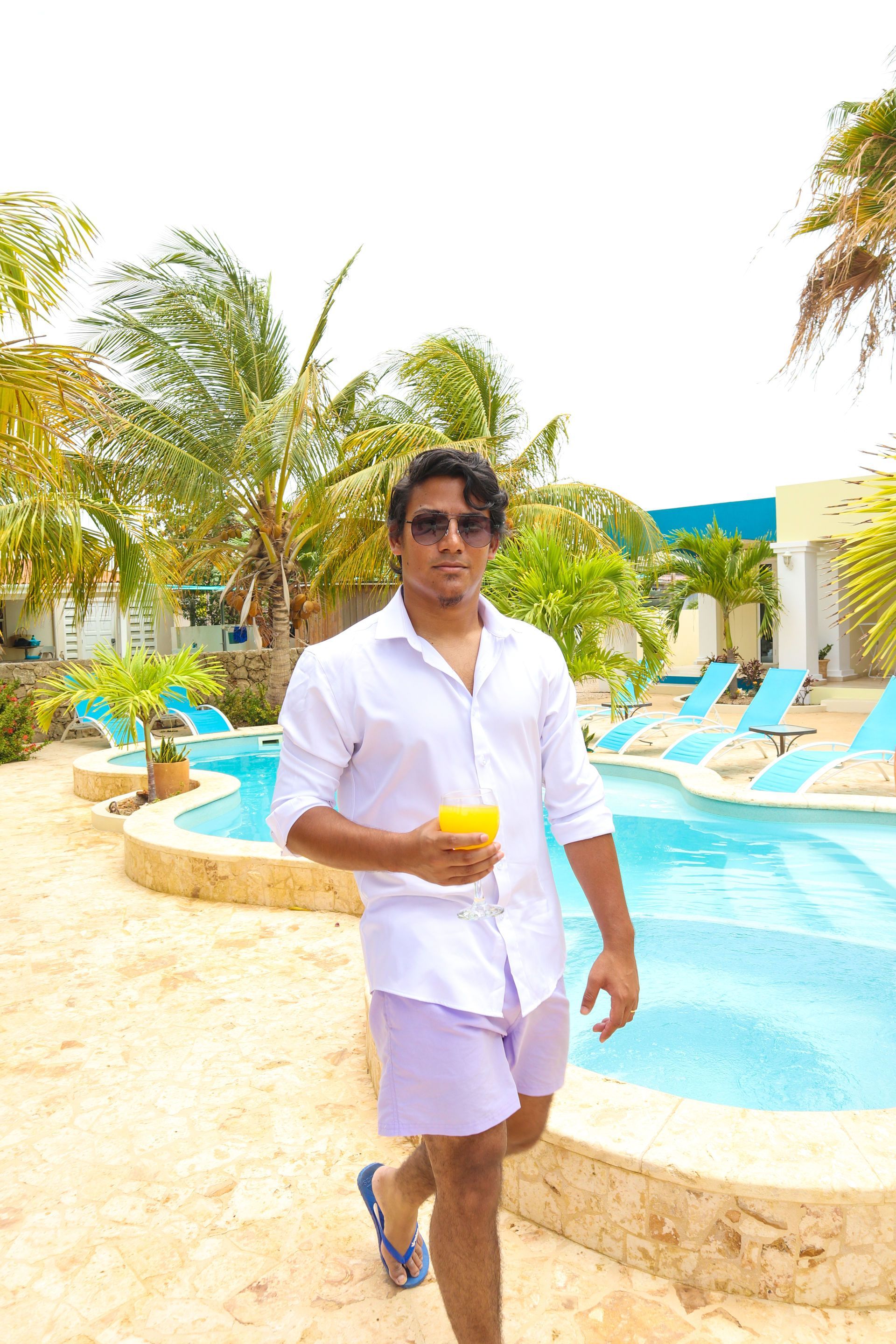 Man in white shirt and purple shorts holding a drink, walking near a pool. Palm trees and blue lounge chairs in background.
