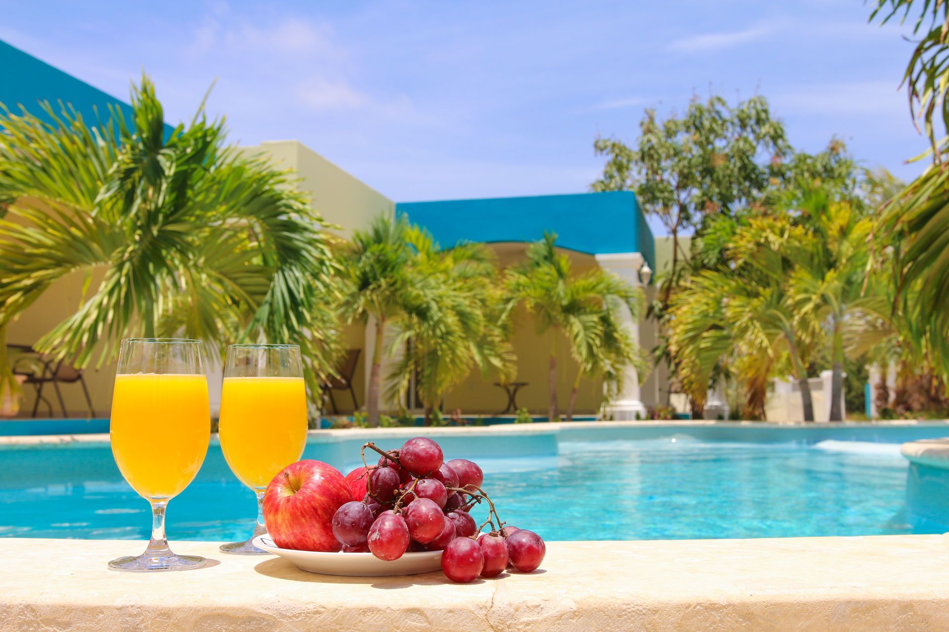 Two glasses of orange juice, grapes, and apples by a pool with palm trees.