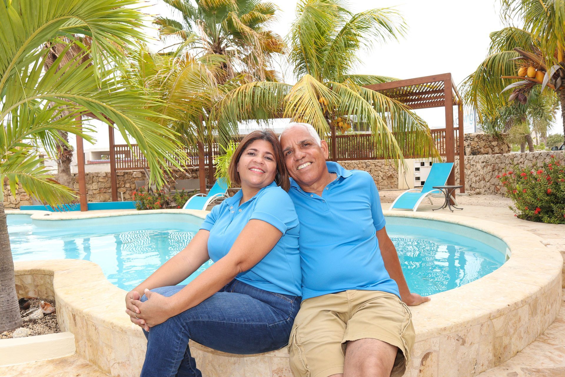 Couple sitting by pool, smiling. Tropical setting with palm trees, blue water, sunny day.