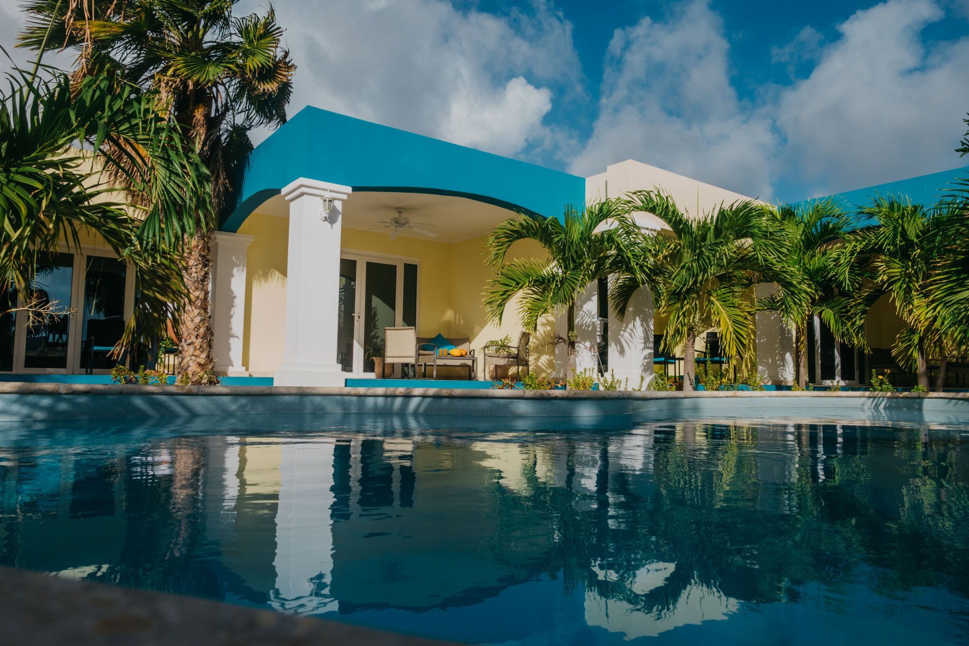 Blue pool reflecting a turquoise-trimmed building with palm trees under a bright sky.