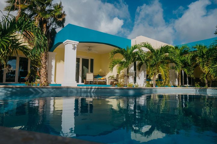 Swimming pool in front of a turquoise and yellow building with palm trees and a cloudy sky.