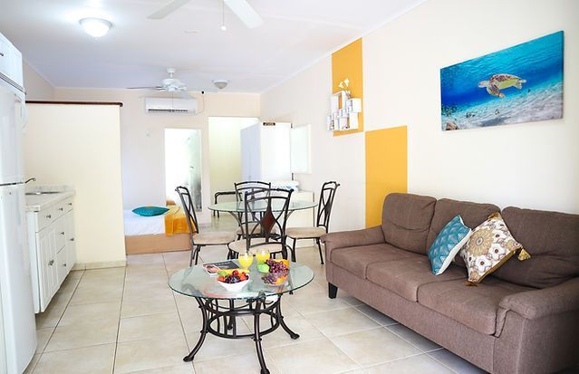 Bright living area with brown sofa, glass table, and dining set; artwork and yellow wall accents.