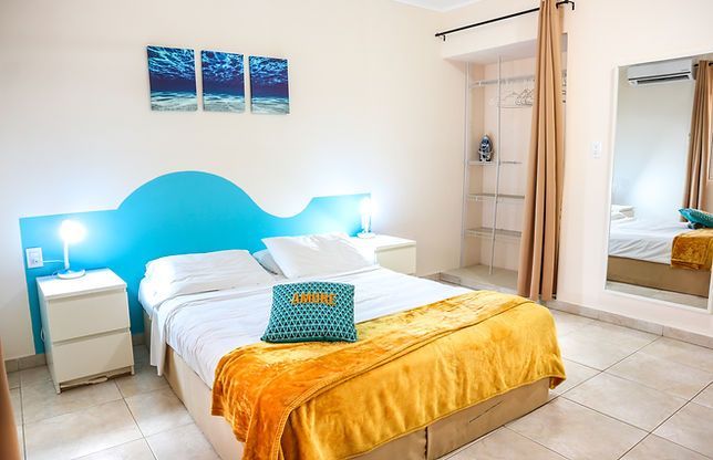 Bedroom with a double bed, light blue headboard, orange blanket, white walls, and a large mirror.