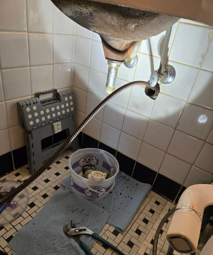 Plumbing work under a sink: a snake, tools, and a bucket are in a tiled bathroom.