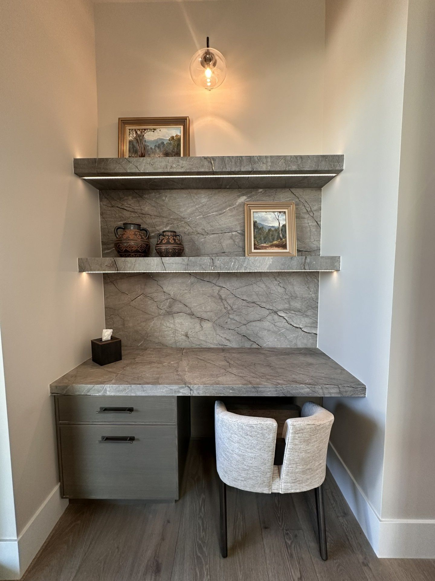 A built-in stone desk and two shelves in a recessed nook with neutral walls, a grey storage cabinet, and a textured chair.