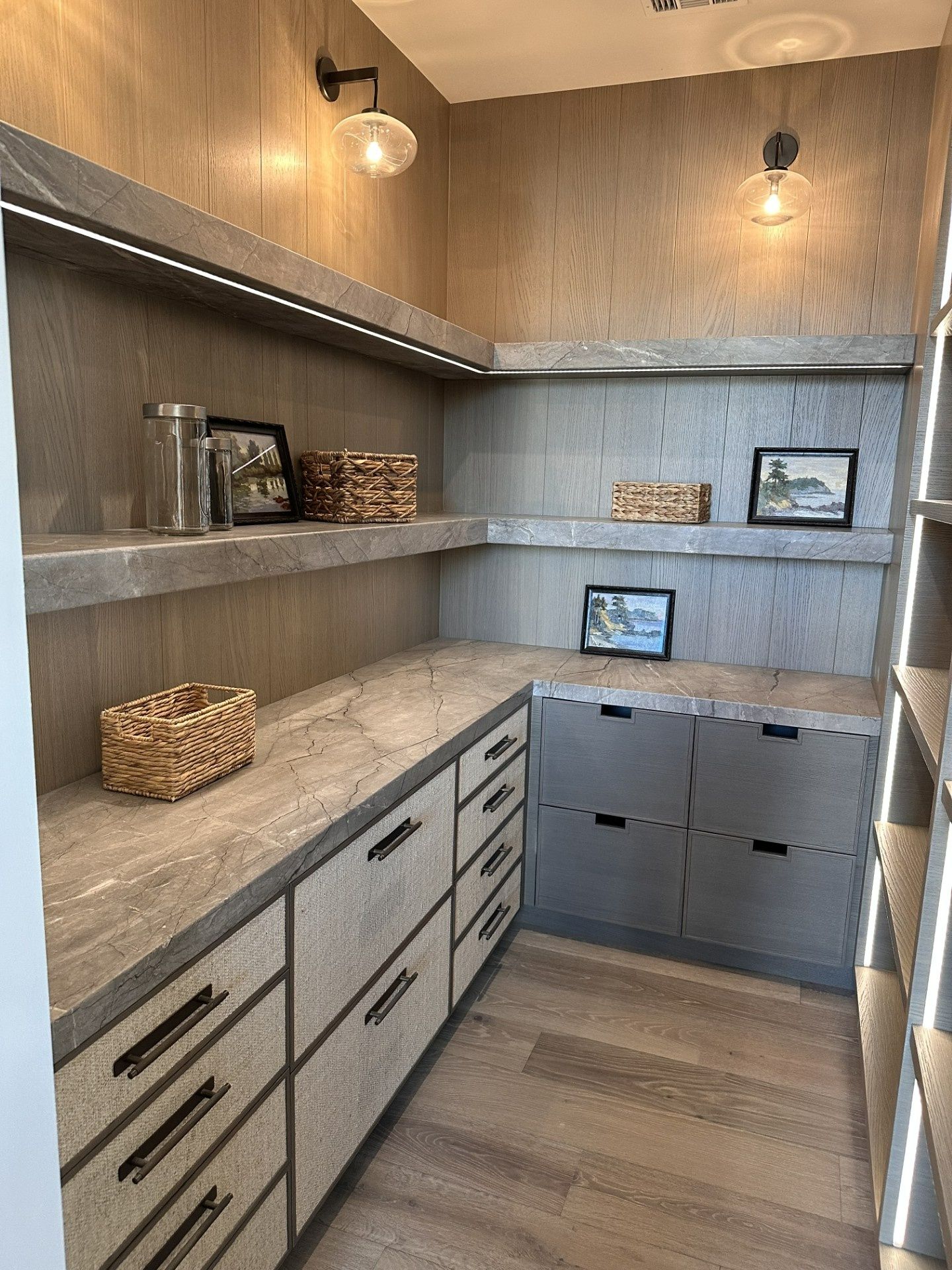 A modern walk-in pantry with light wood cabinetry, stone countertops, open shelving, integrated lighting, and drawers.