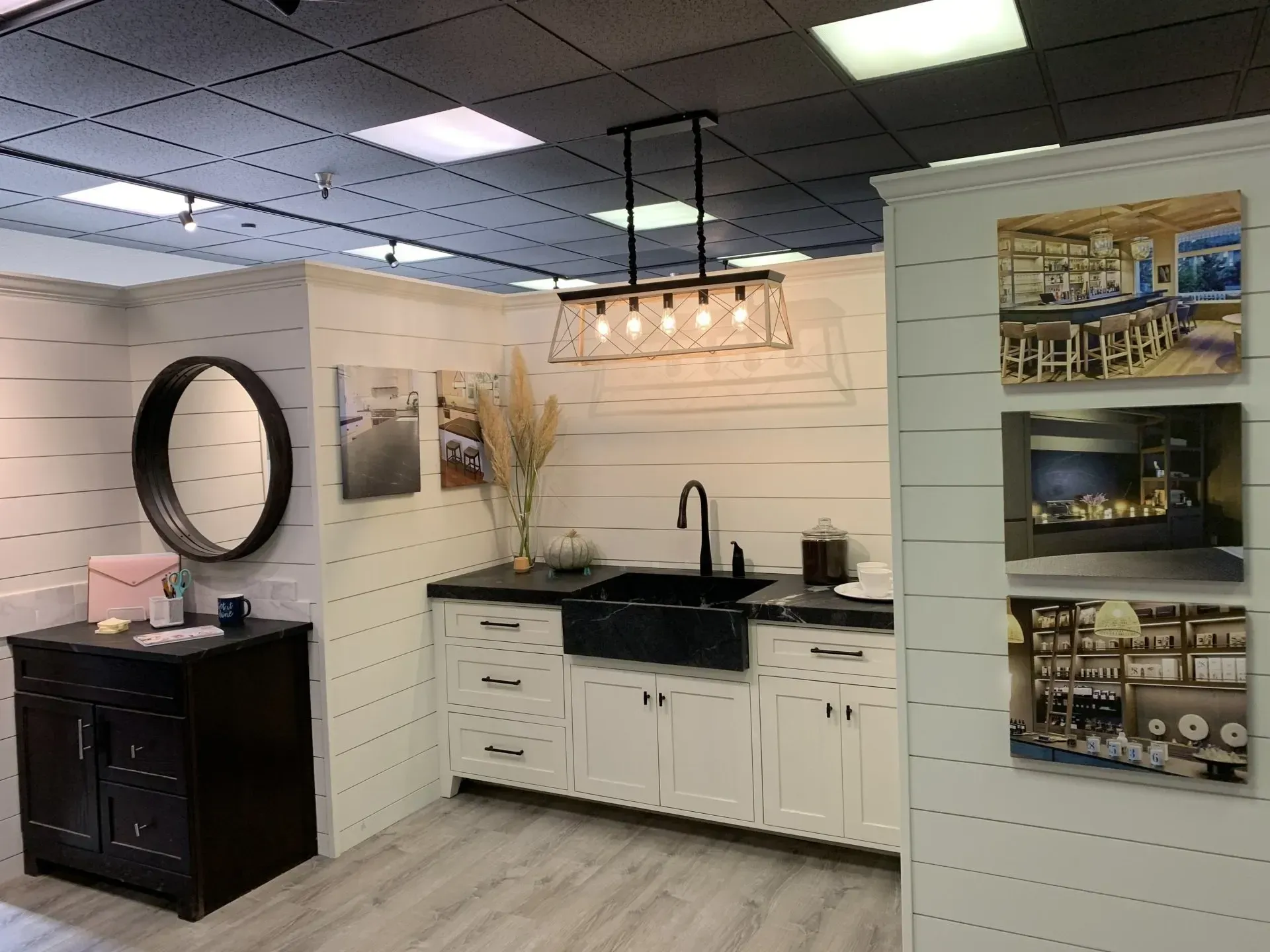 A showroom display featuring a black farmhouse sink, white cabinets, a circular mirror, and hanging light fixtures.