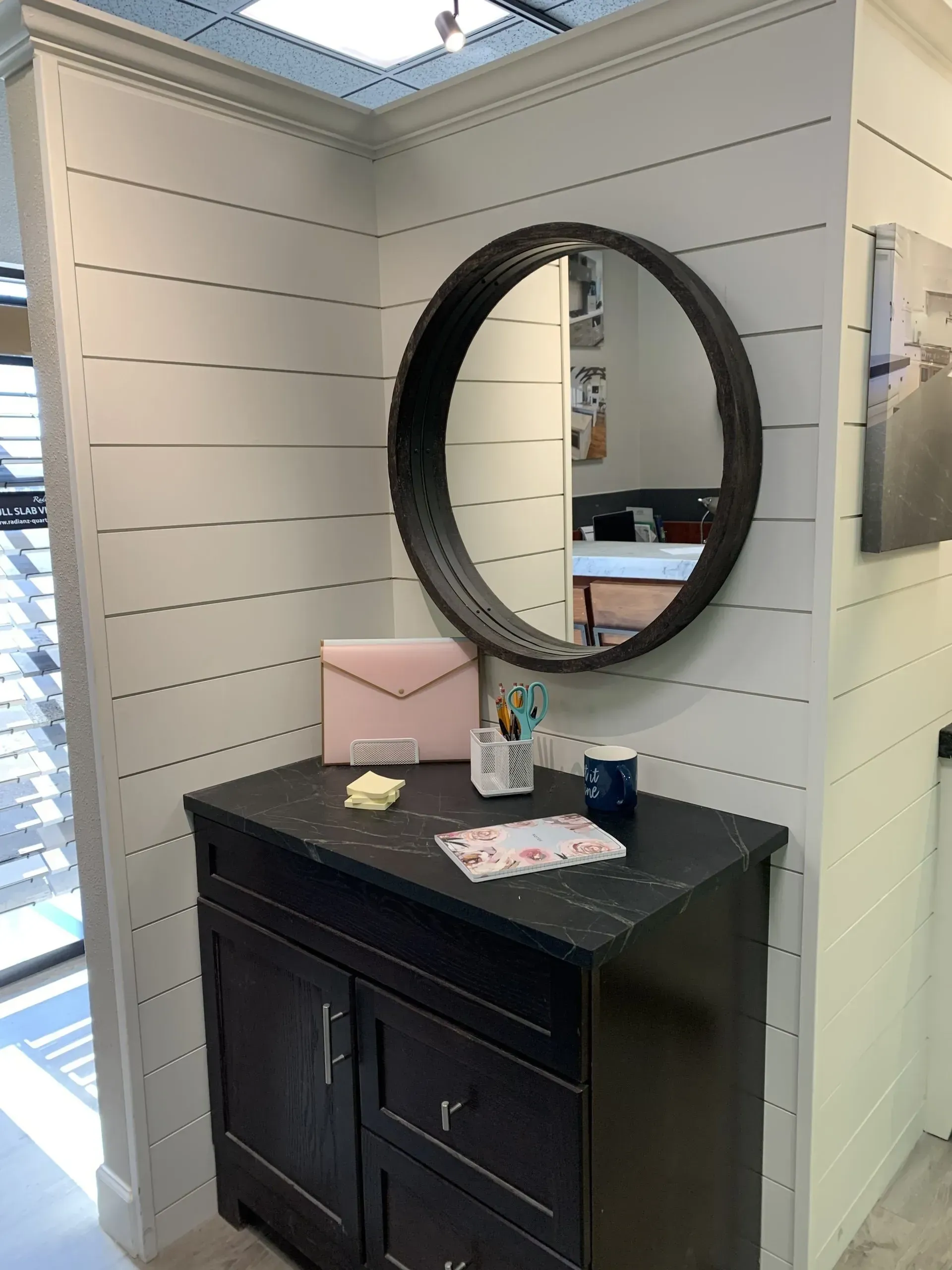 A black wooden cabinet with a round mirror sits against white shiplap walls in a bright, modern entryway.
