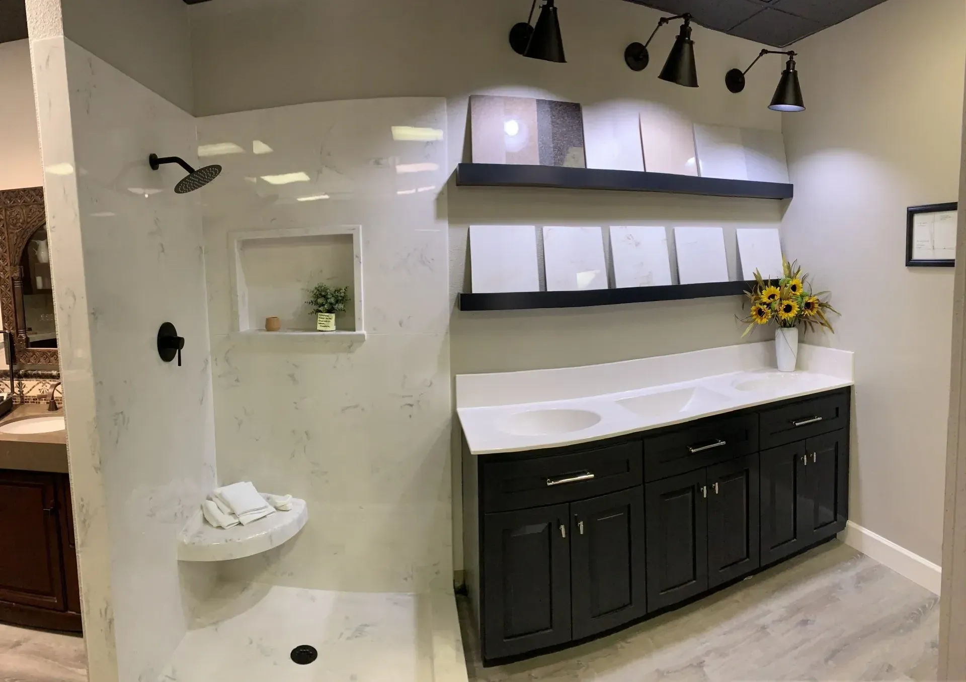 A modern bathroom showroom display featuring a white marble shower, dark cabinets with a white countertop, and wall shelves.