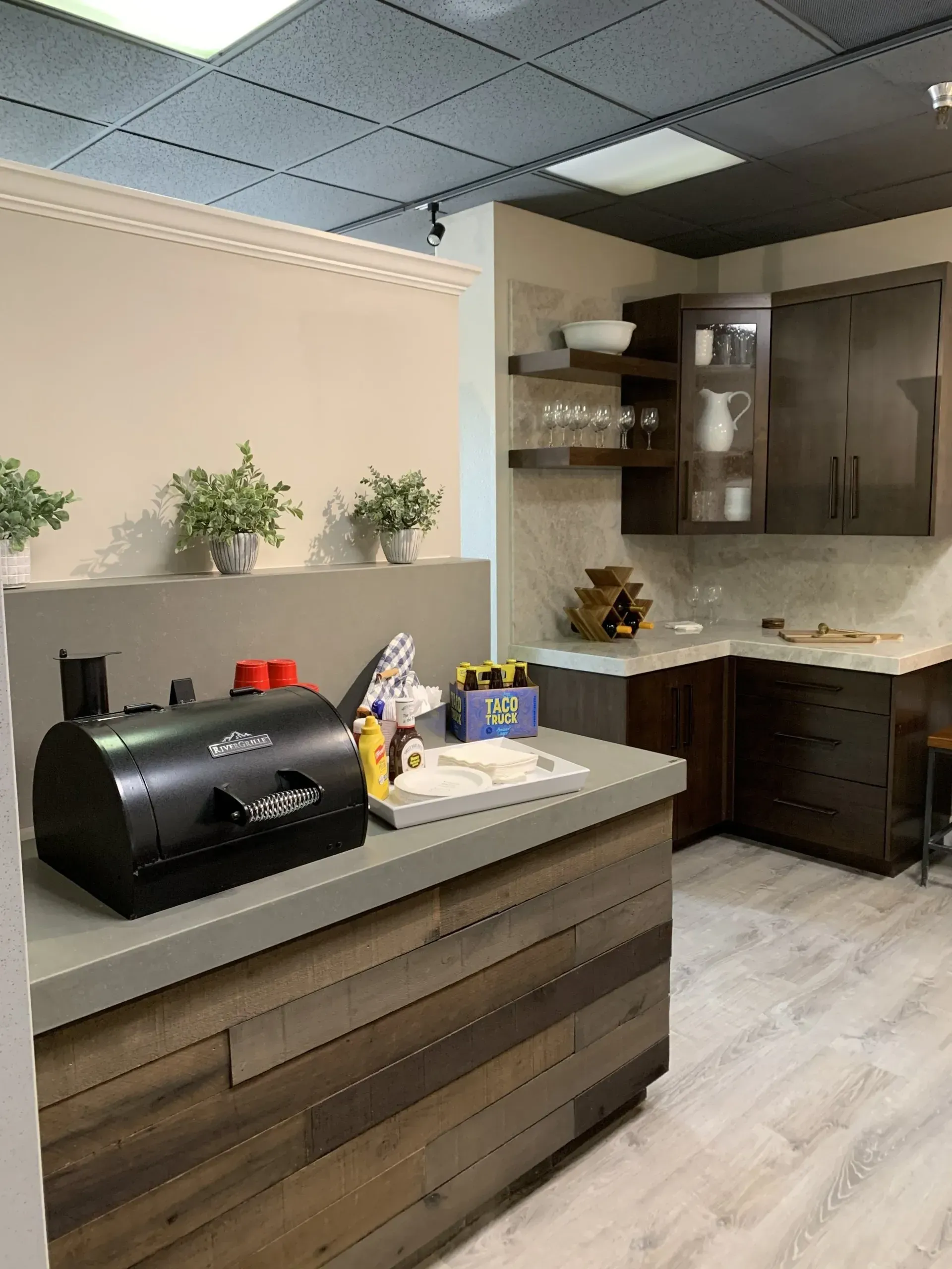 A kitchen corner with a wooden-plank bar, a black grill, small plants, dark cabinets, and light-colored countertops.