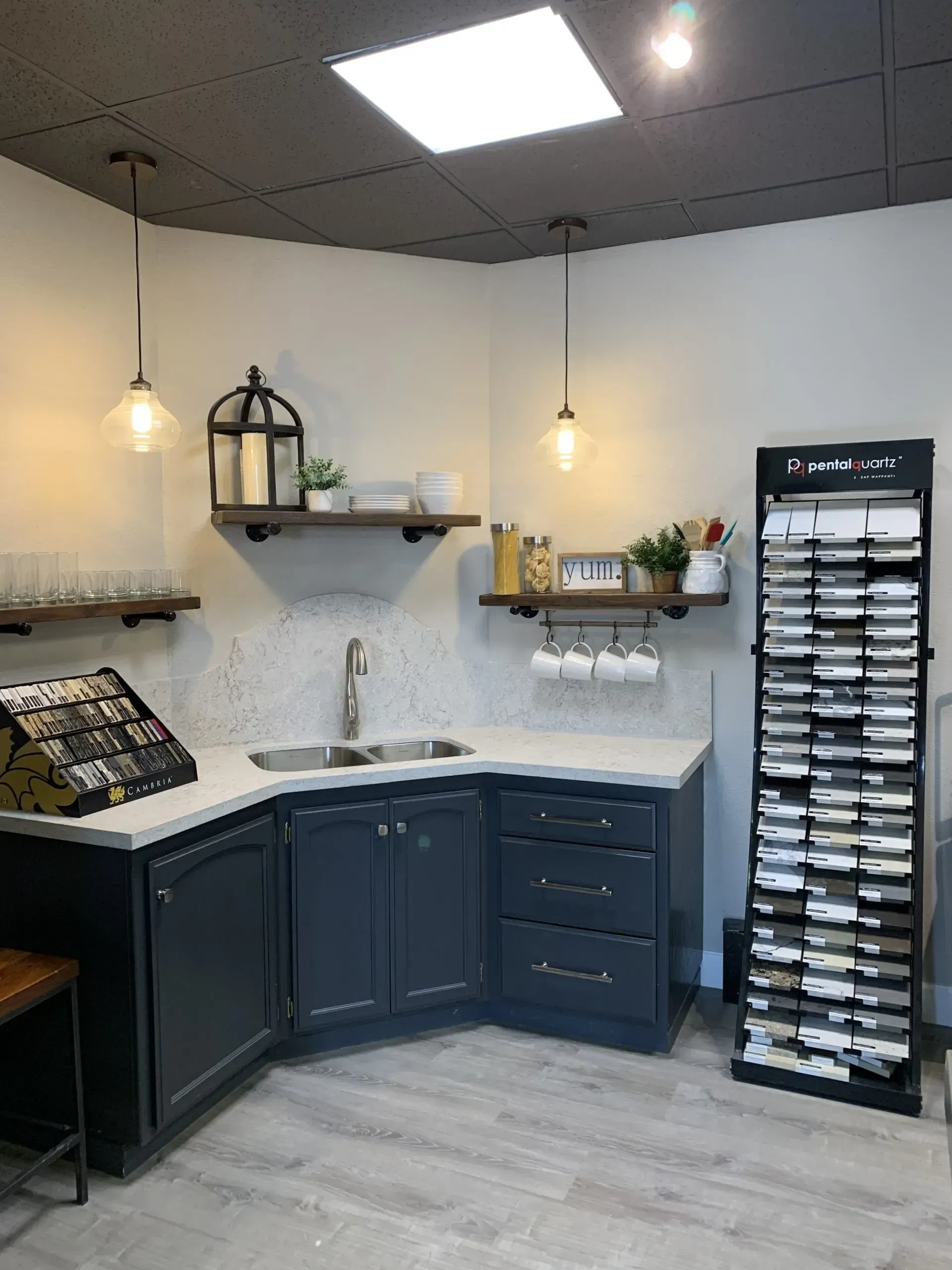 A kitchen showroom corner featuring dark cabinets, a white countertop with a sink, open shelving, and a sample display rack.