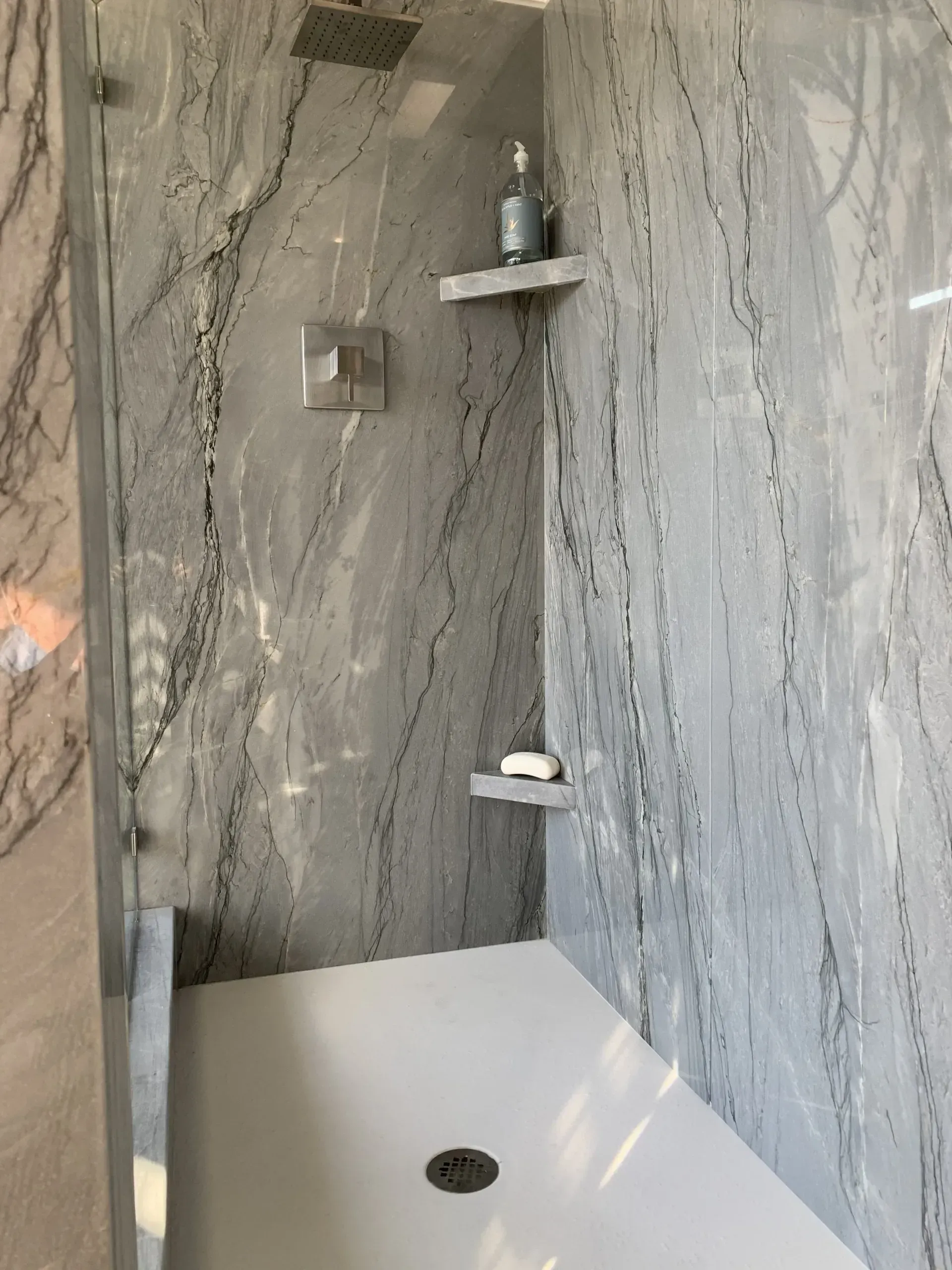 A walk-in shower with gray-veined stone walls, a white floor, chrome fixtures, and two small stone corner shelves.