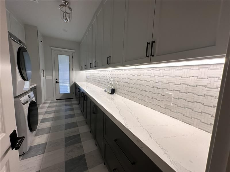 Modern laundry room with stacked washer and dryer, long countertop, dark base cabinets, and white backsplash with LED light.
