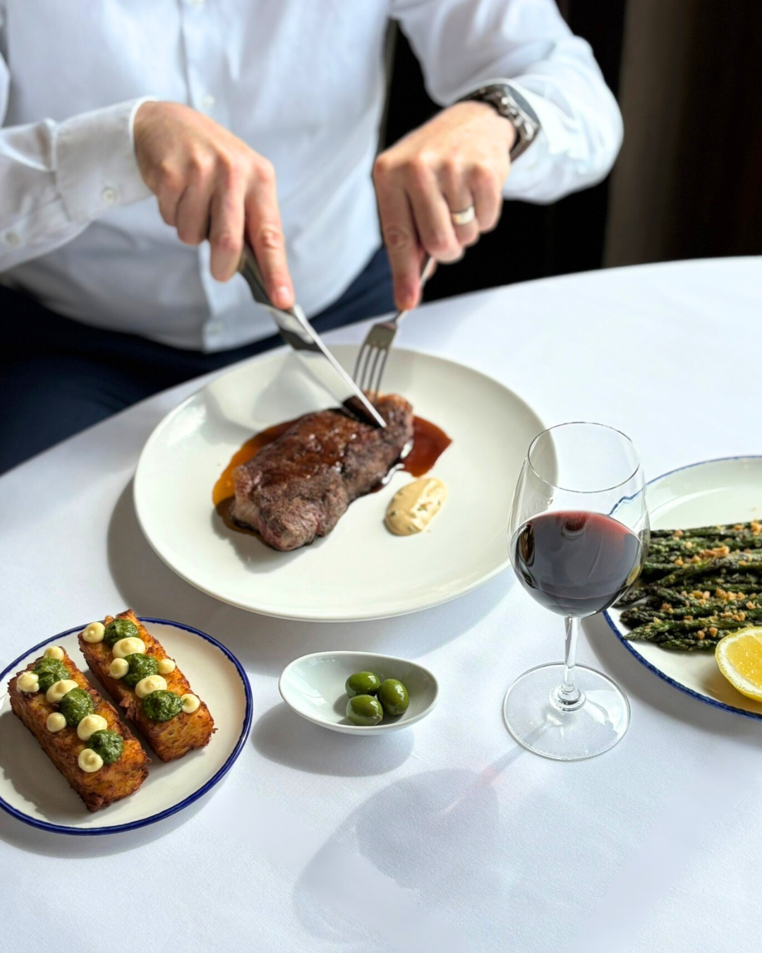Steak being cut on a table covered in a white tablecloth, with wine. 
