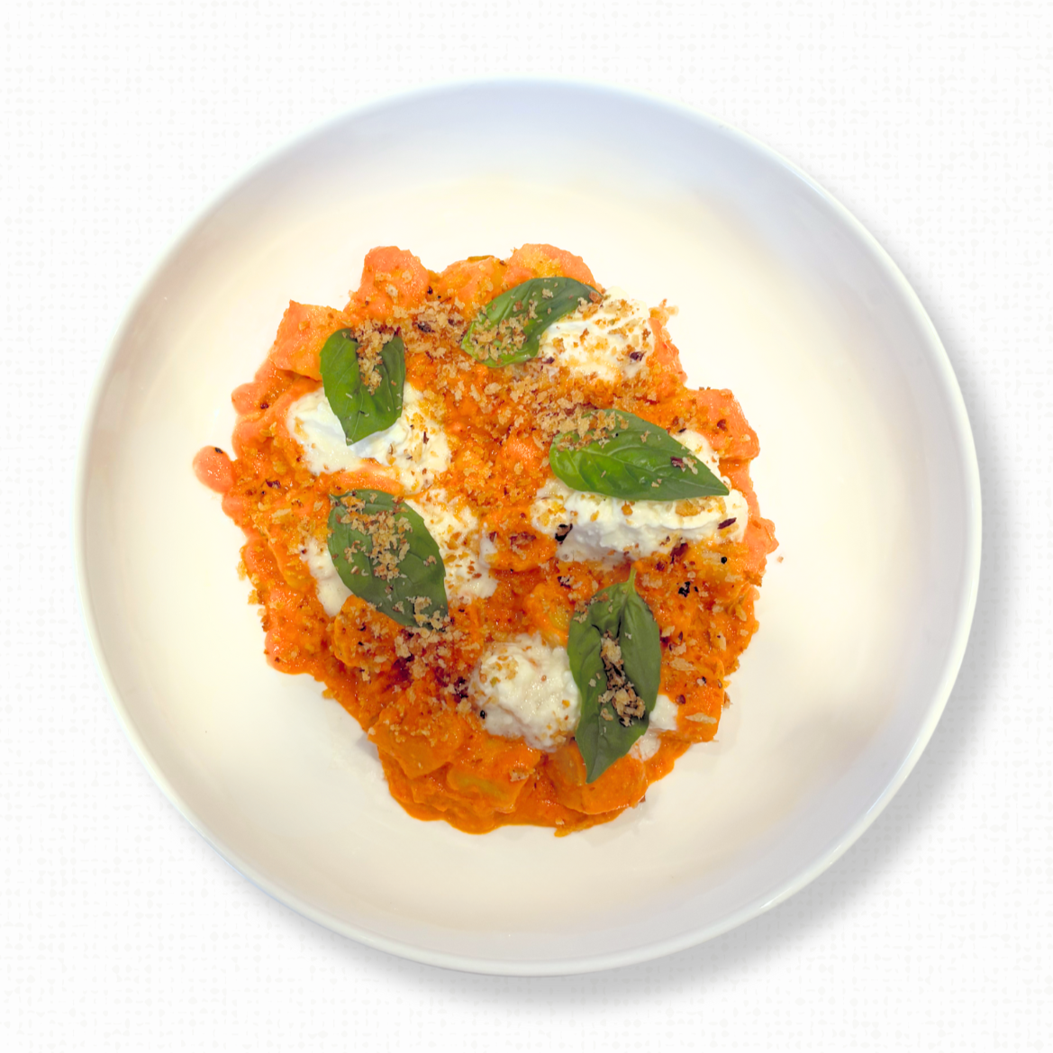 A plate of Gnocchi a la Vodka with a Nduja, Stracciatella, Garlic Breadcrumbs, Parmesan & Fresh Herbs on a white tablecloth-covered table. 